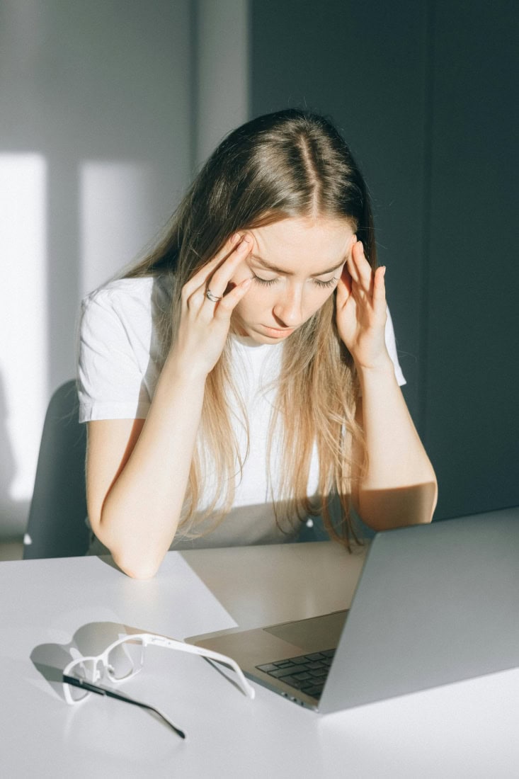Headache due to stress or prolonged work, woman with headache at table with laptop.